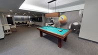 This single-floor amenity space is designed with a sleek, modern aesthetic that caters to both recreation and social gathering. The centerpiece of the room is a full-size pool table with a rich walnut-toned wood frame and vibrant teal felt surface, positioned beneath a rectangular pendant light with a warm bronze shade that draws focus to the playing area. The flooring throughout is a neutral grey carpet that complements the room's cool, contemporary color palette.

A striking oversized billiards-themed mural dominates one wall, depicting a dramatic close-up of pool balls in motion, adding a bold and dynamic visual element to the space. Recessed lighting in the white ceiling provides ambient illumination across the room.

Toward the rear, a casual lounge area features round tables and white chairs, suitable for conversation or spectating. Adjacent to this is a dining nook with bar-height seating and darker furnishings, lending a more intimate feel. A large flat-screen television is mounted on the wall, enhancing the entertainment offering. Together, these zones create a well-rounded, versatile space for leisure and socializing.