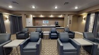 Located at 131 E Bardin Rd in Arlington, Texas, this single-floor private theater room delivers an immersive cinematic experience through a thoughtfully designed layout and premium furnishings. The screening area is anchored by a large wall-mounted flat-screen display, framed by a clean, white built-in surround and flanked by a sleek media console. Rows of plush, dark blue leather recliner chairs with integrated cup holders and side tables are arranged across a patterned, neutral-toned carpet, providing comfortable seating for multiple guests. Dramatic dark drapes line the walls, contributing to the room's light-controlled, theater-like ambiance. Warm wall sconces offer soft, atmospheric lighting throughout the space. At the rear of the room, a full-service refreshment bar features a light-colored countertop, a beverage cooler, a coffee station, and a classic red popcorn machine, ensuring a complete entertainment experience. Recessed ceiling lighting illuminates the bar and lounge areas effectively. The overall design palette of warm taupe walls, deep navy seating, and rich dark accents creates a sophisticated and inviting atmosphere well-suited for private screenings and group gatherings.