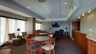 This single-floor lounge space is thoughtfully designed to accommodate both casual gatherings and relaxed individual use. The room features a drop tile ceiling with recessed lighting, which provides consistent, even illumination throughout the space. Subtle textured wallcovering lines the walls, adding depth and a polished, professional character to the interior.

The layout is organized into distinct seating zones. Round dining-height tables paired with cushioned wood-framed chairs offer comfortable spots for small groups, while a separate lounge area provides plush armchairs and a sofa arranged around a wall-mounted flat-screen television — ideal for unwinding or watching programming. Additional accent seating is thoughtfully placed near the windows, which are dressed with valances and sheer curtains that allow soft natural light to filter into the room.

Warm-toned table lamps and a floor lamp complement the overhead lighting, creating an inviting and cozy ambiance. The patterned carpet anchors the space and adds a classic, hospitality-inspired aesthetic. A wooden door with warm-toned millwork is visible along one wall, contributing to the refined overall finish. The space conveys a welcoming, functional atmosphere well-suited for social interaction and relaxation.