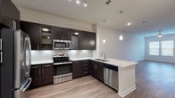 Located at 18610 Tuscany Stone in San Antonio, TX, this single-story residence offers a thoughtfully designed open-concept layout finished with contemporary details throughout. The kitchen serves as the heart of the home, showcasing rich dark cabinetry extending to the ceiling, light granite-style countertops, a subway tile backsplash, and a full suite of stainless steel appliances including a French door refrigerator, electric range, over-the-range microwave, and dishwasher. A peninsula with an undermount sink and pendant lighting above creates a natural transition into the living area. The living space is bright and open, featuring warm wood-style plank flooring, neutral walls, and a window that draws in natural light. The bedroom features plush carpet flooring and a window overlooking outdoor greenery. The primary bathroom is well-appointed with a dual-sink vanity topped with granite-style counters, dark cabinetry, a large framed mirror, and a separate walk-in closet with built-in shelving and hanging space. An additional louvered-door closet provides further storage along the main hallway. The unit's clean, neutral palette and modern finishes create a polished, move-in-ready living environment.