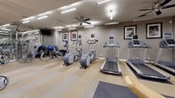 This fitness center at 6355 Peachtree Dunwoody Rd in Sandy Springs, Georgia, offers a thoroughly outfitted, single-floor exercise facility designed to accommodate a wide variety of workout routines. The space is characterized by neutral gray walls, patterned carpet flooring with rubber mat zones, and a bright, well-lit environment achieved through a combination of overhead fluorescent lighting and ceiling fans that contribute to a comfortable workout atmosphere.

The cardio section features multiple treadmills and elliptical machines, each equipped with individual display screens, positioned along one wall beneath a row of framed athletic artwork that adds an energizing aesthetic to the space. The strength training area is expansive, housing a variety of cable machines, a power rack with adjustable bench, a full dumbbell rack, and multiple selectorized weight machines suited for both upper and lower body training. Stability balls and additional accessories are also available for functional fitness exercises.

Floor-to-ceiling mirrors line key areas of the facility, visually expanding the space and allowing users to monitor their form throughout workouts. Ceiling fans are distributed throughout, enhancing air circulation. The facility also includes a clearly marked exit corridor with an adjacent glass-door room, as well as hygiene stations for user convenience. The overall design is clean, functional, and professional.
