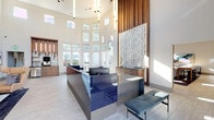 Located at 4407 Hopson Rd in Morrisville, NC, this contemporary single-floor clubhouse is thoughtfully designed to serve as a dynamic social and professional hub. The grand entrance lobby immediately impresses with soaring ceilings, dramatic vertical pendant lighting, and a sleek, high-gloss blue reception desk set against a striking textured stone accent wall framed in warm wood paneling. Floor-to-ceiling windows flood the space with an abundance of natural light and offer views of the surrounding community. A bright sunroom-style lounge area features expansive multi-pane windows, navy blue accent chairs, and an open, airy atmosphere. Enclosed private workspaces with glass-paneled doors provide quiet areas for focused work, while a cozy banquette nook and casual seating areas with warm amber chairs offer inviting spots for conversation. The game room is well-appointed with a billiards table, shuffleboard, wall-mounted displays, and stylish wallpaper accents. A fully equipped kitchen and social bar area showcases a marble waterfall island, dramatic clustered pendant lighting, and a navy tile feature wall with built-in media display. Throughout, light wood-tone flooring, curated contemporary artwork, and a cohesive blue-and-neutral palette create a polished, welcoming ambiance.