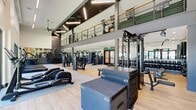 This two-story fitness center at 3099 Iowa Ave in Riverside, CA totals 2,350 square feet, with 1,510 sq ft on the ground floor and 840 sq ft on the upper level. The facility features soaring double-height ceilings in key areas, large floor-to-ceiling windows, and sleek wood-look flooring throughout, creating a bright and modern atmosphere. The ground floor houses an extensive array of equipment, including cable machines, a functional trainer rig with battle ropes and a heavy bag, a full dumbbell rack with adjustable benches, and Life Fitness strength machines. A striking geometric-patterned accent wall anchors the main strength area, adding a bold design element. Cardio equipment, including treadmills and ellipticals, is positioned along expansive windows offering views of an outdoor turf training area, accessible via glass double doors. Ceiling fans and roller shades provide climate and light control. The upper level, accessible via an open staircase with metal railings, features spin bikes, resistance training equipment, and mirror-lined walls. A green accent wall and textured tile detailing add visual contrast. The mezzanine overlooks the main floor below, reinforcing the facility's open, connected layout. The overall design is modern and industrial, combining functionality with high-end aesthetic finishes.