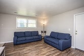 This single-story home offers a straightforward, functional layout designed for comfortable everyday living. The living room serves as the central gathering space, featuring rich wood-look flooring that extends throughout the main areas and neutral gray walls that lend a clean, understated aesthetic. Two navy blue sofas anchor the room, and a window along one wall brings in natural light with views of surrounding greenery, creating an inviting atmosphere.

The floor plan, as revealed by the aerial view, connects the living area to a dining space and kitchen in an efficient arrangement. The kitchen is equipped with warm oak cabinetry offering ample storage both above and below the countertops, a double-basin sink, white appliances including a refrigerator and range, and a microwave. Light tile flooring keeps the space bright and easy to maintain.

The bedroom is a well-proportioned retreat featuring the same wood-look flooring, a ceiling fan with light fixture, and a wide window that allows generous natural light to fill the room. The rustic barn-style headboard adds a touch of character to the neutral space.

The bathroom is efficiently designed with tile flooring, a vanity with wood cabinetry, and a shower-tub combination enclosed by a curtain, providing all essential amenities in a compact, functional layout.