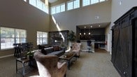 This community room offers an inviting and well-appointed space designed for residents to gather, relax, and socialize. The room immediately impresses with its dramatic double-height ceilings, which are lined with a generous row of clerestory windows that flood the interior with abundant natural light throughout the day. Warm, neutral-toned walls complement the rich textures found throughout the space.

A striking stone fireplace serves as the focal centerpiece of the main lounge area, adding a cozy and refined architectural element. The seating arrangement features a plush gray sofa and complementary upholstered armchairs surrounding a wood-and-metal coffee table, creating an intimate yet open gathering area. Patterned carpeting anchors the lounge zone, while hard flooring extends toward the adjacent areas.

Toward the back of the room, a well-equipped kitchenette area is finished with dark cabinetry, a light-toned countertop bar, and pendant lighting that adds a stylish accent. A refrigerator and workspace are visible, making the area functional for events and casual use. Potted greenery is thoughtfully placed throughout the room, adding a natural, lively touch to the overall ambiance. The space strikes a balance between comfort and functionality, making it an ideal hub for community activity.