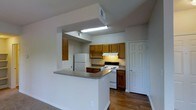 This single-floor space showcases a thoughtfully designed kitchen that serves as the heart of the layout. The kitchen is defined by warm, medium-toned wood cabinetry that lines the walls, offering generous storage through both upper and lower cabinet configurations. A peninsula counter with a laminate surface extends into the adjacent living area, creating a natural divide between spaces while also providing additional workspace and informal seating potential.

The kitchen is equipped with essential appliances, including a white refrigerator and a white electric range with an overhead vent hood, both neatly integrated into the cabinetry arrangement. Recessed overhead fluorescent lighting illuminates the cooking area efficiently, complemented by the under-cabinet light above the range. The flooring transitions from hardwood-style material in the kitchen zone to carpeting in the adjoining living area, subtly delineating each functional space.

White painted walls and light-toned ceilings contribute to a clean, neutral aesthetic throughout. A paneled white door on the far wall leads to an additional room, and a visible HVAC vent in the ceiling suggests consistent climate control. Overall, the space is practical and straightforward in its design.