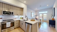 This thoughtfully designed one-bedroom, one-bathroom residence offers a cohesive modern aesthetic throughout its efficiently planned single-floor layout. The open-concept kitchen is a standout feature, showcasing wood-grain flat-front cabinetry extending to the ceiling, a classic subway tile backsplash, stainless steel appliances including a full-size refrigerator, range, microwave, and dishwasher, and crisp white quartz countertops. A peninsula with seating for multiple bar stools connects the kitchen seamlessly to the living area, making it an ideal space for both cooking and entertaining. Light wood-tone flooring flows continuously throughout, lending warmth and visual cohesion. The living area benefits from direct access to an outdoor space, with glass doors that invite natural light deep into the unit. The bedroom is a calm and comfortable retreat, featuring multiple windows with exterior views, a ceiling fan, and ample room for a large bed and additional furnishings. A dedicated closet provides practical storage. The bathroom features a glass-enclosed walk-in shower with large-format tile, matte black hardware accents, and light wood flooring that continues the home's clean design language. White panel doors with matte black hardware are used throughout, maintaining a consistent and polished modern style.