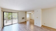 This single-floor unit is characterized by a clean, open layout with light wood-style flooring running throughout, creating a cohesive and airy feel across all areas. The living space is generously proportioned, divided into two interconnected zones — a front area with direct access to an outdoor patio through a sliding glass door fitted with vertical blinds, and a second living or dining area equipped with a ceiling fan and a window that brings in natural light. A wall-mounted air conditioning unit provides climate control in the main living zone.

The kitchen is efficiently designed and features white cabinetry with warm wood-toned trim, granite-style countertops, a double stainless steel sink positioned beneath a window with outdoor views, and standard appliances including an electric stovetop and dishwasher. The kitchen opens toward the dining area, supporting a connected flow between cooking and living spaces.

A mirrored sliding-door closet is visible near one of the main rooms, offering practical storage. The bathroom includes a tub and shower combination enclosed by sliding frosted glass doors with chrome framing. Throughout the unit, neutral white walls and consistent flooring contribute to a light, understated, and functional ambiance.