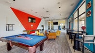 Rebel Retreat is a lively, single-floor community clubhouse located in Oxford, Mississippi, designed to foster social interaction and entertainment. The space makes a bold visual statement with its striking color palette — vivid red geometric accent walls, teal-painted surfaces, and crisp white ceilings — creating an energetic yet cohesive atmosphere throughout.

The main gathering area serves as the heart of the clubhouse, featuring wood-look plank flooring and an open layout that accommodates multiple activity zones. A full-size billiards table and a shuffleboard table anchor the game area, while wall-mounted flat-screen TVs provide entertainment from multiple vantage points. Comfortable lounge seating, including tan leather chairs and bold yellow sofas, offers inviting spots to relax between games.

A dedicated bar and kitchen area extends the social experience, outfitted with a long countertop bar, plush blue velvet barstools, dark wood cabinetry with frosted glass accents, and stylish Sputnik-inspired pendant lighting. A foosball table adds another recreational option in this area, and large windows flood the room with natural light while providing views of the surrounding greenery.

The perimeter of the clubhouse features floor-to-ceiling black-framed glass windows and doors that open to an exterior covered patio, seamlessly blending indoor and outdoor spaces. High-top tables and bar seating along the teal accent wall create an additional casual dining and social zone. Decorative wall art and ceiling fans complete the well-appointed interior.
