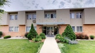 This residential building presents a classic and tidy exterior characterized by a combination of warm brick and neutral lap siding that gives it a timeless, approachable aesthetic. The main entrance is framed by a covered awning supported by a clean architectural overhang, flanked by traditional wall-mounted lantern-style sconces that add a welcoming touch. A straight, well-maintained concrete walkway leads visitors directly to the glass-paneled entryway, creating a clear and accessible path to the front door. The building number is prominently displayed near the entrance for easy identification. The grounds surrounding the building are thoughtfully landscaped, featuring a variety of mature evergreen shrubs, ornamental trees, and seasonal flowering plants that add color and vibrancy to the façade. A well-kept lawn stretches across the front, contributing to the overall curb appeal. The building spans two stories, with large windows on both levels allowing for natural light throughout the interior units. The upper level features a balcony-style railing, adding architectural interest to the exterior profile. Overall, the building exudes a sense of established, well-cared-for residential living in a community-oriented setting.