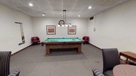 This single-floor recreational space is thoughtfully designed to serve as a social and leisure hub, offering two distinct activity zones that flow naturally into one another. The main area is anchored by a full-size pool table with a rich wood frame and vibrant green felt surface, illuminated by an elegant pendant light fixture that incorporates a decorative clock — a functional and stylish centerpiece. Framed artwork adorns the neutral-toned walls, while a pair of deep red leather armchairs provide comfortable seating for spectators. The carpeted flooring adds warmth throughout the space, complementing the neutral wall tones and drop tile ceiling fitted with recessed lighting. A wall-mounted cue rack keeps equipment neatly organized and accessible. Adjacent to the billiards area, a separate game room zone is accessible through a classic wood-framed French door, lending an air of distinction between the two spaces. This secondary area features a round wooden table surrounded by striped upholstered chairs, set up with a chess board — ideal for card games, board games, or casual conversation. A traditional table lamp on a side console adds a warm, inviting glow. Together, the two zones create a well-appointed recreational environment suited for group gatherings and friendly competition.