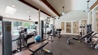 This fitness center is a thoughtfully designed workout facility that combines functional amenities with an open, airy atmosphere. The space is defined by its striking vaulted ceiling accented with exposed dark wood beams, which lend a warm architectural character to the room. Pendant lights and ceiling fans are suspended overhead, complementing the abundant natural light that floods in through expansive floor-to-ceiling windows and glass doors along the perimeter walls. These windows offer views of lush, tree-lined outdoor surroundings and a glimpse of a pool area, enhancing the overall ambiance of the space.

The floor is covered in durable dark rubber matting, well-suited for heavy equipment and high-traffic use. The fitness center is comprehensively equipped for both strength and cardiovascular training. Strength equipment includes a multi-station cable machine, a functional trainer, weight benches, a full dumbbell rack with a wide range of free weights, and various selectorized resistance machines targeting different muscle groups. Cardio offerings include a Precor treadmill, an upright cycle, and additional aerobic equipment positioned near the windowed walls.

A separate adjoining room, accessible through glass-paned French doors, appears to serve as a secondary fitness or wellness space. The overall environment is bright, spacious, and well-organized, providing a complete and inviting setting for a thorough workout.