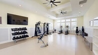 Located at 8300 Merrill Rd in Jacksonville, FL, this fitness center offers a spacious, single-floor layout designed to support a comprehensive workout experience. The room features elevated tray ceilings accented by large ceiling fans, which contribute to both the airy ambiance and effective air circulation throughout the space. Soft, neutral yellow-toned walls paired with classic white wainscoting lend the facility a clean, polished aesthetic. The flooring is a smooth, light-toned surface that is easy to maintain and comfortable underfoot.

Natural light floods the space through an expansive bank of large, multi-pane windows along one wall, offering views of the surrounding greenery and keeping the environment bright and inviting. A wall-mounted flat-screen television is positioned for visibility across the workout area, adding an entertainment element to exercise sessions.

The fitness center is well-appointed with a diverse array of equipment, including a treadmill, rowing machine, elliptical trainers, a multi-station cable and weight machine, a dedicated free weight rack with dumbbells, and adjustable workout benches. Additional accessories and mats are neatly stored along the walls. A set of glass-paneled French doors provides a refined entry point, completing the upscale yet functional character of this well-designed fitness facility.