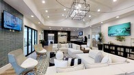 This single-floor space makes an immediate impression with its soaring vaulted ceilings and striking geometric chandelier centerpiece, crafted in a dark iron-and-glass cage style that anchors the room with both drama and elegance. Recessed lighting throughout supplements the chandelier, ensuring the space is well-lit and inviting at any time of day.

The main lounge area is generously proportioned, featuring an expansive arrangement of crisp white sofas and accent chairs layered over a bold monochromatic patterned area rug. Black and white throw pillows and sculptural décor accessories add visual interest while maintaining a cohesive, refined color palette. Woven rattan accent chairs introduce a touch of natural texture, softening the overall look.

Toward the rear, a dark-toned wet bar and kitchenette area provides a functional counterpoint to the light-filled lounge, complete with built-in shelving and a wall-mounted television. Adjacent to this is a dining or café-style seating zone, suggesting the space is designed for both relaxed socializing and casual dining.

Large glass doors and windows along the perimeter flood the interior with natural light and offer glimpses of lush greenery outside, creating a seamless connection between indoors and out. The overall ambiance is polished, modern, and welcoming.