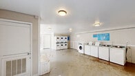 Located at 1025 S Crater Rd in Petersburg, Virginia, this single-level community laundry facility offers a spacious, open layout designed to accommodate multiple users simultaneously. The room features white-painted walls accented by wainscoting panels along the lower half, lending a clean and utilitarian aesthetic throughout the space.

The laundry area is well-equipped with a row of top-loading washing machines lined along one wall, accompanied by a front-loading washer, providing ample capacity for resident use. Stacked dryer units are positioned in an adjacent section, maximizing the use of vertical space. Informational signage is posted near the machines for user guidance.

Flush-mounted ceiling light fixtures provide consistent overhead illumination throughout the facility. The concrete floor spans the entirety of the room, reflecting light from a glass-panel exterior door that allows natural light to filter in and provides outdoor access. A mailbox station is also visible within the space, suggesting this facility serves as a shared common area for a residential community.

An interior door with a no-smoking sign connects to an additional room or corridor, and a low built-in bench near the entrance offers a convenient spot for residents. The open floor plan ensures easy movement and functionality throughout this practical, community-oriented space.