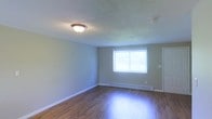 This two-story residential space presents a straightforward and functional design with practical finishes throughout. The interior features warm hardwood flooring that adds character and durability underfoot, complemented by crisp white baseboards that frame each room with a clean, finished look. Walls are painted in a neutral, soft tone that creates a calm and welcoming atmosphere, providing a versatile backdrop for any décor style. The textured popcorn ceilings are fitted with flush-mounted light fixtures, ensuring consistent and even illumination throughout the space. A standout feature of the main living area is the baseboard heating system, which runs along the lower perimeter of the walls, offering efficient and evenly distributed warmth. Natural light enters through a sliding window dressed with vertical blinds, brightening the interior while allowing for adjustable privacy and light control. The overall layout across both floors provides ample space for comfortable living, with a practical design that prioritizes functionality. The neutral palette and classic finishes make this space a solid canvas that is move-in ready and easy to personalize to a variety of tastes and lifestyles.