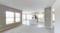 This single-floor residence showcases an open-concept layout that integrates the living area and kitchen into one cohesive, light-filled space. The living area features plush carpeting and is lined with multiple windows dressed with white blinds, allowing abundant natural light to pour in and create an airy, welcoming atmosphere. Neutral greige walls complement the clean white trim and baseboards throughout, lending the space a modern yet timeless aesthetic. The transition from carpet to large-format tile marks the entry into the kitchen zone, where a central island with a crisp white countertop serves as both a functional workspace and a natural gathering point. The kitchen is well-appointed with white shaker-style cabinetry, stainless steel appliances including a French door refrigerator with an ice and water dispenser, and a dishwasher. A dedicated sink area sits beneath a window, filling the workspace with natural light. Track lighting above the kitchen ensures ample task illumination. The overall design emphasizes openness and functionality, with a neutral palette and clean finishes that provide a versatile backdrop for a variety of interior styles.