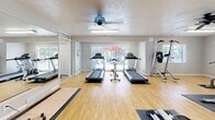 Located in Roy, Utah, this fitness center offers a thoughtfully organized exercise space designed to support a wide variety of workout routines. The room features smooth, light wood flooring throughout, with rubber mat zones strategically placed beneath equipment to provide stability and protection during workouts.

The space is comprehensively outfitted with both cardio and strength training equipment. Cardio options include multiple treadmills, a stationary bike, an elliptical machine, and a rowing machine, providing ample variety for aerobic exercise. On the strength side, the room houses a multi-function cable machine, a dedicated weight bench, a full rack of dumbbells ranging across multiple weights, a medicine ball storage tower, and an inversion table, among other equipment.

Large mirrors line one wall, enhancing the sense of openness while allowing users to monitor their form. Sliding glass doors connect the fitness room to an adjacent interior space and allow natural light to filter in, while multiple windows along the exterior walls offer views of the surrounding outdoor amenity area, including a glimpse of a pool. Dual ceiling fans and fluorescent lighting ensure the space remains comfortable and well-lit during any workout. The textured ceiling adds a subtle architectural detail to the otherwise clean, functional interior.