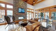 This single-story community clubhouse blends rustic craftsmanship with modern comfort through a cohesive design language of warm wood tones, natural stone, and expansive glazing. The grand entrance foyer welcomes visitors with soaring vaulted ceilings supported by exposed timber trusses, wide-plank hardwood floors, and floor-to-ceiling wood-framed windows that flood the space with natural light and offer views of the surrounding outdoors. A dedicated gallery-style hallway features curated botanical artwork and a console table, providing an elegant transition between areas. The centerpiece of the main lounge is a dramatic floor-to-ceiling stacked stone fireplace, topped by a mounted flat-screen television and flanked by large French doors that open directly to an outdoor patio. Comfortable leather sofas and an area rug anchor the seating arrangement, creating an inviting gathering space. Adjacent to the lounge, a well-lit fitness area is visible through glass partitions, housing cardio equipment in a bright, open setting. A separate conference or meeting room, accessible via wood-framed glass doors, provides a functional space for group activities. Throughout the clubhouse, consistent use of timber detailing, warm neutral tones, and thoughtful lighting — including a statement chandelier — reinforces a refined yet welcoming mountain-lodge aesthetic.