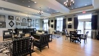This sophisticated single-floor space exudes a timeless, refined ambiance through a carefully curated black-and-white design palette. The open layout is anchored by tray ceilings adorned with ornate chandelier lighting, lending a sense of grandeur and vertical interest to the room. Warm hardwood flooring flows throughout, providing an inviting contrast to the bold furnishings. Plush black leather sofas and patterned armchairs are arranged to encourage comfortable conversation and relaxation, while coordinating black-and-white checkered drapery frames the large windows that allow abundant natural light to fill the space. Wall sconces and elegant floor lamps supplement the ambient lighting, creating a warm and welcoming atmosphere at any hour. A curated gallery of framed artwork and decorative mirrors adorns the walls, enhancing the classic aesthetic. A dining area with dark wood chairs and tables is positioned near the windows, offering a bright and stylish setting for gatherings. The overall design strikes a balance between formal elegance and everyday comfort, making this a versatile space well-suited for both quiet retreats and social occasions.