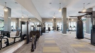 This expansive fitness center offers a generously proportioned, single-level layout designed to accommodate a comprehensive range of workout routines. The space is defined by soaring ceilings, pendant lighting with an industrial-chic aesthetic, and large picture windows that flood the interior with natural light while providing views of an outdoor pool and surrounding community. The flooring features a patterned carpet tile design in neutral tones of gray, tan, and gold that delineates workout zones throughout the space. Full-length mirrors line entire walls, visually amplifying the already generous footprint and allowing users to monitor their form during exercise. The cardio zone is well-equipped with multiple treadmills, ellipticals, and stair climbers positioned directly in front of the oversized windows. The strength training area features a variety of cable machines, multi-function weight stations, selectorized machines, a dumbbell rack, flat benches, and a heavy punching bag. A dedicated functional training zone includes resistance bands and TRX-style equipment. A striking large-scale mural depicting Texas Longhorn cattle on an open pasture serves as a bold focal point, adding local character and visual interest. Wall-mounted televisions and ceiling fans complete this thoughtfully appointed fitness facility.