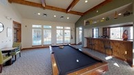 This single-floor community or clubhouse space is thoughtfully designed to accommodate both relaxation and recreation. The centerpiece is a spacious game room featuring soaring vaulted ceilings with exposed wooden beam accents, adding a warm, rustic character to the space. A full-size pool table anchors the room, which is bathed in natural light from a generous bank of large windows overlooking an outdoor area. Adjacent to the game room is a built-in wet bar with a raised countertop and bar seating, ideal for entertaining. A dedicated activity nook provides a chess table and comfortable seating beneath the high ceilings, while a wall-mounted clock adds a classic touch. Connected lounge areas offer plush leather seating and access to additional rooms through wide, wood-trimmed doorways. The fully equipped kitchen features warm honey-toned cabinetry, stone tile flooring, stainless steel appliances including a refrigerator, electric range, and over-the-range microwave, as well as stone countertops. Patterned carpeting flows throughout the main gathering areas, complementing the natural wood trim and neutral wall tones. The overall ambiance is inviting and functional, well-suited for community gatherings or social events.