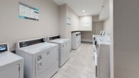 This shared laundry facility at 115 Hutchinson Ave in Columbus, Ohio, offers a clean and efficiently organized space designed to serve multiple users with ease. The room is finished in neutral warm-gray tones with smooth walls and light-colored large-format tile flooring, contributing to a bright, hygienic atmosphere. Recessed ceiling lighting provides consistent illumination throughout the space. Multiple front-load and top-load washing machines are arranged along both sides of the room, paired with a corresponding set of dryers, giving users ample options for completing laundry efficiently. Each machine features a digital control panel, and instructional signage is posted prominently on the walls, covering topics such as best practices, reporting laundry issues, and payment guidance. A built-in counter shelf area on one side provides a convenient folding or staging surface. The room also includes a door leading to an adjacent corridor, ensuring easy access from the main building. The layout is thoughtfully arranged to allow comfortable movement between machines, making the facility practical and user-friendly. Overall, the space reflects a modern, no-frills utility design that prioritizes functionality and cleanliness for everyday use.