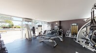 This fitness center is a thoughtfully designed, single-level space that offers a comprehensive workout environment. The layout is divided into distinct functional zones, allowing for a smooth flow between cardio and strength training areas. One side of the facility is lined with floor-to-ceiling glass windows and sliding doors that flood the space with natural light and provide open views of the surrounding outdoor area, including palm trees and landscaped greenery, creating an airy and motivating atmosphere.

The cardio zone is equipped with multiple treadmills, an elliptical cross-trainer, and a stair climber, positioned to take advantage of the natural light and outdoor views. A wall-mounted television is also present for added convenience during workouts. The strength training area features a variety of resistance machines, including cable systems and multi-use weight stations, complemented by a well-stocked dumbbell rack with kettlebells. Adjustable weight benches are positioned throughout for versatile training options.

Dark charcoal walls contrast with the bright, smooth white ceiling, giving the space a modern and focused aesthetic. The floors are finished with durable rubber matting, ensuring safety and practicality. Climate control is provided via wall-mounted air conditioning units. A mirrored wall in the strength area enhances the sense of space and allows users to monitor their form effectively.