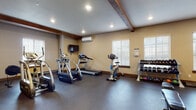 This fitness center at StoneBridge offers a comprehensive workout environment designed to accommodate a wide range of exercise routines. The space is defined by its warm, rustic-modern aesthetic, featuring exposed wood ceiling beams, recessed lighting, and neutral-toned walls that create an inviting and focused atmosphere. Durable dark rubber flooring spans the main exercise area, providing traction and cushioning underfoot.

The cardio zone is equipped with a treadmill, elliptical trainer, stair climber, and stationary bike, all positioned to take advantage of natural light streaming in through multiple windows fitted with white blinds. A wall-mounted flat-screen television accompanies the cardio equipment, offering entertainment during workouts. A wall-mounted mini-split HVAC unit ensures comfortable climate control year-round.

The strength training area includes a multi-tiered dumbbell rack stocked with a full range of free weights, kettlebells, medicine balls, and a stability ball. An adjustable weight bench and suspension training straps mounted from the ceiling provide versatile options for functional fitness. An open cubby shelving unit holds additional fitness supplies. A separate alcove with carpeted flooring houses an additional strength machine, offering a quieter, more focused training space. French glass doors connect the fitness center to adjacent areas, enhancing the open, accessible feel of the overall facility.