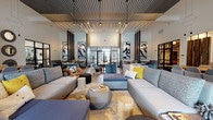 This single-floor clubhouse is a well-appointed communal space designed with a modern, upscale aesthetic throughout. The main lounge area is the centerpiece of the space, showcasing soaring ceilings with a distinctive slatted ceiling detail and large floor-to-ceiling windows that invite abundant natural light and offer views of the surrounding outdoor areas. The flooring features a striking herringbone wood-look pattern that flows seamlessly across the space, anchoring the various seating zones. Generous sectional sofas, accent chairs, and sculptural side tables are thoughtfully arranged to encourage both social gatherings and relaxed lounging, complemented by bold abstract wall art and decorative longhorn accents that add a touch of regional character. A coffee and beverage bar area is equipped with a coffee machine, wine refrigerator, and a communal high-top table with bar seating, set against a dramatic herringbone wood accent wall. Multiple distinct seating vignettes, defined by cowhide and patterned area rugs, provide intimate conversation areas. Glass-paneled partitions offer transparent separation between zones, including a view into an adjacent fitness center. The overall ambiance is contemporary and inviting, balancing rich textures, curated décor, and functional amenities throughout.