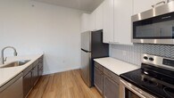 Located at 3900 W Broad St in Richmond, Virginia, this modern single-floor residence offers a thoughtfully designed open-concept layout with clean lines and contemporary finishes throughout. The expansive living area is bathed in natural light through multiple large windows with blinds, showcasing warm hardwood-style flooring that flows seamlessly through the main living spaces. The well-appointed kitchen features two-tone cabinetry — crisp white upper cabinets paired with darker lower cabinets — complemented by a herringbone tile backsplash, stainless steel appliances including a refrigerator, electric range, and built-in microwave, and a light-colored countertop island with teal cabinetry accents. A stacked in-unit washer and dryer is conveniently tucked within a dedicated closet near the living area. The bedrooms are well-proportioned, with hardwood-style flooring and bright windows offering natural light. A generously sized walk-in closet with built-in wire shelving provides ample storage. The unit includes two full bathrooms, each featuring a soaking tub with shower combination, brushed nickel fixtures, grab bars, and tile flooring. The overall ambiance is bright, clean, and move-in ready, reflecting modern residential design.