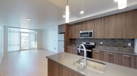 This single-floor space showcases a contemporary open-concept layout that seamlessly connects the kitchen and living area. The kitchen is well-appointed with flat-front cabinetry finished in a warm wood-grain tone, complemented by a grey tile backsplash that adds texture and visual interest. Stainless steel appliances, including a refrigerator, electric range, and over-the-range microwave, provide a polished, functional finish. A spacious island with a quartz-style countertop and an integrated sink serves as a central focal point, ideal for both meal preparation and casual dining. A contemporary pendant light hangs above, adding a refined design touch. Light wood-tone flooring runs throughout, contributing to the bright, airy atmosphere of the space. Recessed lighting in the ceiling ensures even illumination across the kitchen and living areas. The adjoining living space benefits from natural light filtering in through floor-to-ceiling windows dressed with vertical blinds, enhancing the overall sense of openness. Clean white walls and neutral tones throughout create a cohesive, modern aesthetic.