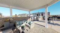 This single-floor residence offers a thoughtfully designed layout that balances comfort and functionality. The open-concept kitchen is equipped with stainless steel appliances, including a refrigerator and microwave, complemented by dark cabinetry and granite-style countertops. A breakfast bar with seating for two provides a casual dining option and connects seamlessly to the main living areas. Crown molding throughout adds a refined architectural detail, while neutral-toned walls and plush carpeting in the living spaces create a warm, inviting ambiance. The living room is generously sized, featuring a large window that allows natural light to fill the space, and offers ample room for a full seating arrangement. An arched hallway leads to additional rooms, maintaining an elegant flow throughout the unit. A dedicated display area adds a decorative touch, ideal for showcasing personal accents. The outdoor covered patio is a standout feature, finished with a cobblestone floor and enclosed by a stone knee wall with black iron railing. It provides a relaxing lounge space with wicker seating and open views of the surrounding residential community under a bright, open sky.