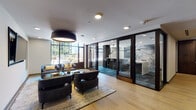 Located at 198 SW Columbia St in Portland, Oregon, this contemporary single-floor space is thoughtfully designed to offer a range of distinct functional zones within a cohesive, upscale aesthetic. Light-toned hardwood flooring flows throughout much of the space, complemented by recessed lighting and bold pendant fixtures that anchor each seating area.

The main lounge area features plush dark leather armchairs and sofas arranged around low coffee tables atop a woven area rug. Floor-to-ceiling windows along one wall flood the space with natural light and provide views of the surrounding urban environment. A separate lounge zone offers navy upholstered seating grouped around a central table, flanked by built-in wall niches displaying curated reading materials and a wall-mounted flat-screen television.

A standout bar area features a generous marble-topped counter with modern swivel bar stools. Behind the counter, a striking large-scale fingerprint graphic serves as a focal point against dark tile and wood-paneled walls, illuminated by a sleek linear pendant light.

A corridor lined with intimate booth seating, separated by striped wood-and-fabric dividers set against deep teal accent walls, provides semi-private nooks. Glass-paneled doors connect the various zones, maintaining visual openness while defining each distinct area.