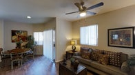 This single-story space showcases a thoughtfully designed living area that balances comfort and functionality. The room features rich hardwood-style flooring that runs throughout, adding warmth and visual continuity to the space. Neutral beige walls provide a versatile backdrop, complementing the warm, earthy tones of the furnishings.

A central ceiling fan with an integrated light fixture serves as a practical focal point overhead, ensuring year-round comfort and ambient lighting. Two windows dressed with vertical blinds allow for adjustable natural light, brightening the room while maintaining privacy. A sliding glass door leads to an adjoining outdoor area, further enhancing the sense of openness and providing additional natural light.

The living area accommodates a plush sofa adorned with patterned accent pillows, paired with a wicker-style coffee table that adds a natural texture to the space. A side table with a table lamp contributes to the layered lighting scheme, creating a cozy atmosphere. Decorative elements, including indoor plants and ceramic accents, complete the inviting, homey character of this well-appointed living space.