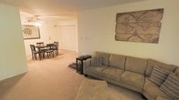 This single-floor space showcases an open-concept layout that seamlessly connects the living and dining areas, creating a cohesive and functional environment. The living area is anchored by a neutral-toned sofa complemented by a tufted ottoman and a compact side table, while a decorative world map wall piece serves as a notable focal point, adding character to the space. The walls are finished in a soft, warm neutral tone that enhances the overall inviting ambiance. Plush wall-to-wall carpeting in a warm beige hue runs throughout, contributing to a comfortable, understated aesthetic. The dining area, visible from the living space, accommodates a dark wood table and chair set, making it well-suited for everyday meals or casual gatherings. A set of large closet doors can be seen in the background, offering practical storage solutions. The open flow between the living and dining zones maximizes the usable space while maintaining an airy, uncluttered feel. Overhead lighting provides consistent illumination throughout, ensuring both areas are well-lit and functional.