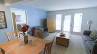 This single-floor space offers a welcoming and functional layout that seamlessly blends the dining and living areas into one cohesive environment. The interior is immediately distinguished by its calming blue accent walls, which contrast pleasantly with the neutral grey tones of the carpeted flooring throughout.

The dining area is furnished with a warm wood-toned round table and matching chairs, providing an inviting spot for meals or casual gatherings. A decorative floral arrangement adds a vibrant pop of color to the space. Flowing naturally from the dining area, the living space accommodates a comfortable grey upholstered sofa accented with blue throw pillows, alongside a substantial wood armoire that provides generous storage.

Natural light plays a key role in the ambiance of this space, with multiple large windows and a glass-paneled exterior door flooding the interior with brightness and creating an airy, open feel. The door also offers the potential for outdoor access, enhancing the sense of connectivity between the interior and exterior.

The overall design is clean and practical, with a neutral palette punctuated by warm wood furnishings and thoughtful décor details. The open-plan arrangement maximizes the sense of space, making the layout feel expansive and easy to navigate.