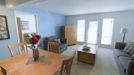 This single-floor space offers a welcoming and functional layout that seamlessly blends the dining and living areas into one cohesive environment. The interior is immediately distinguished by its calming blue accent walls, which contrast pleasantly with the neutral grey tones of the carpeted flooring throughout.

The dining area is furnished with a warm wood-toned round table and matching chairs, providing an inviting spot for meals or casual gatherings. A decorative floral arrangement adds a vibrant pop of color to the space. Flowing naturally from the dining area, the living space accommodates a comfortable grey upholstered sofa accented with blue throw pillows, alongside a substantial wood armoire that provides generous storage.

Natural light plays a key role in the ambiance of this space, with multiple large windows and a glass-paneled exterior door flooding the interior with brightness and creating an airy, open feel. The door also offers the potential for outdoor access, enhancing the sense of connectivity between the interior and exterior.

The overall design is clean and practical, with a neutral palette punctuated by warm wood furnishings and thoughtful décor details. The open-plan arrangement maximizes the sense of space, making the layout feel expansive and easy to navigate.