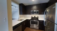 This single-floor space showcases a well-appointed kitchen designed with a sharp contrast between dark espresso cabinetry and crisp white countertops, creating a bold, contemporary aesthetic. The kitchen is equipped with a full suite of stainless steel appliances, including an electric range with a double oven, an over-the-range microwave, a dishwasher, and a refrigerator. The cabinetry extends to the ceiling, providing generous upper and lower storage throughout. A white subway tile backsplash adds a clean, classic touch that complements the modern design palette. The undermount single-basin sink is paired with a sleek gooseneck faucet, positioned conveniently along the counter. Hardwood flooring runs through the space, adding warmth that balances the kitchen's cooler tones. A glimpse into an adjacent area reveals recessed track lighting, suggesting a connected living or utility space. Overall, the kitchen exudes a functional yet stylish character, with quality finishes and a thoughtfully organized layout suited for everyday cooking and entertaining.
