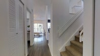 This two-story home offers a thoughtfully designed interior that balances simplicity with comfort. The main level is anchored by rich, dark hardwood floors that run throughout, creating a warm contrast against the bright white walls and trim. A central hallway connects the various living areas seamlessly, with an open sightline leading toward the main living space, where large sliding glass doors or floor-to-ceiling windows invite abundant natural light and provide views of a lush outdoor balcony or patio area. The living area feels airy and spacious, enhanced by the generous flow of light that fills the room. A staircase with a textured woven carpet runner and clean white railings leads to the upper floor, adding a classic yet understated design element. The hallway also features convenient storage with louvered closet doors, contributing to the home's organized and functional layout. The overall ambiance throughout the space is calm and inviting, with a neutral palette of whites and natural wood tones creating a timeless, move-in-ready aesthetic. The combination of open sightlines, natural light, and cohesive finishes gives the home a polished, well-maintained character across both levels.