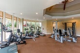 Located at 580 Arastradero Rd in Palo Alto, CA, this fitness center offers a thoughtfully designed single-floor workout facility. The space is defined by its striking octagonal architecture, highlighted by a raised, coffered ceiling with exposed wooden beams that adds both height and visual character to the room. Recessed lighting throughout ensures a bright, well-lit environment, while a checkerboard-patterned carpet floor provides comfortable, durable underfoot support.

Floor-to-ceiling glass windows and sliding glass doors wrap generously around the space, flooding the interior with natural light and offering direct views of the surrounding lush landscaping and an outdoor pool area — creating an inviting, open atmosphere during every workout.

The facility is comprehensively equipped for a full range of training needs. Cardio equipment includes treadmills with integrated screens, stationary bikes, ellipticals, and a recumbent bike. A dedicated strength training area features a multi-station cable machine, adjustable weight benches, kettlebells, medicine balls, and resistance accessories neatly organized on wall-mounted racks. A large wall mirror enhances the strength training zone, adding depth to the space. The combination of natural light, greenery views, and varied equipment makes this fitness center a functional and pleasant environment for residents.