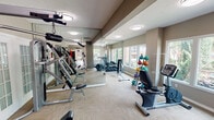 Located at 6601 Treepoint Dr in Arlington, Texas, this fitness center is a well-equipped, single-floor facility designed to support a full range of workout routines. The space is organized into distinct zones, seamlessly connected by carpeted flooring and neutral, warm-toned walls throughout.

The cardio area features multiple treadmills, elliptical machines, and a stationary recumbent bike, all positioned near large windows that invite natural light and offer views of the surrounding outdoor greenery. The strength training section is well-appointed with cable machines, multi-functional weight stations, an adjustable bench, a dumbbell rack, and a colorful selection of medicine balls and kettlebells. Full-length mirrors with glass partitions line one side of the space, enhancing the sense of openness and allowing users to monitor their form.

French doors with glass panes provide access to an adjacent outdoor area, where seating is visible, adding a casual relaxation option. A Coca-Cola vending machine is conveniently situated near the entrance to this outdoor connection. Flush-mount ceiling lights provide consistent, even illumination throughout, complementing the natural light from the windows. The textured ceiling and structured column accents contribute to the architectural character of the space, making this fitness center both functional and inviting.