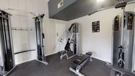 This fitness center is a fully outfitted, single-floor facility designed to support a comprehensive workout routine. The space is divided into distinct functional zones, seamlessly integrated within an open layout. The cardio area lines one side of the room, housing multiple Spirit-brand treadmills, an elliptical trainer, and an upright cycle, all positioned beside large windows that offer views of an outdoor pool and surrounding landscaping, flooding the area with natural light. The strength training zone features a dual-cable functional trainer, a multi-station cable machine, a lat pulldown unit, and an incline bench, providing a thorough range of resistance training options. Instructional exercise posters are mounted on the walls for user reference. The interior is finished with white vertical shiplap-style paneling on the lower walls, contrasted by a dark upper wall treatment and ceiling, lending a modern, structured aesthetic. Durable dark rubber flooring covers the entire space, offering both safety and sound absorption. Multiple entry and exit points are accessible, including a door with windows that opens directly to the outdoor pool area, enhancing the sense of connection to the exterior environment.