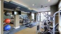 This fitness center offers a comprehensive workout environment thoughtfully designed for a range of exercise needs. The space is organized into distinct zones, with a dedicated free weights area featuring a full dumbbell rack and exercise mats, alongside a multi-station cable machine and additional strength training equipment positioned throughout the floor. Cardio machines, including treadmills and ellipticals, are arranged along one section, each equipped with individual monitors to keep users engaged during workouts. Structural columns with dark accent detailing define the layout, adding an architectural dimension to the space while also helping to segment workout zones. The flooring combines neutral carpeting with interlocking rubberized tiles in the weight area, providing both comfort and durability. A wall-mounted flat-screen television enhances the cardio area, while ceiling fans promote air circulation throughout. Large windows along the far wall invite ample natural light, creating an open and airy atmosphere. Mirrored walls further contribute to the sense of spaciousness. Overall, the fitness center presents a clean, modern environment well-suited for a full-body workout routine.