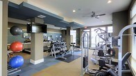 This fitness center offers a comprehensive workout environment thoughtfully designed for a range of exercise needs. The space is organized into distinct zones, with a dedicated free weights area featuring a full dumbbell rack and exercise mats, alongside a multi-station cable machine and additional strength training equipment positioned throughout the floor. Cardio machines, including treadmills and ellipticals, are arranged along one section, each equipped with individual monitors to keep users engaged during workouts. Structural columns with dark accent detailing define the layout, adding an architectural dimension to the space while also helping to segment workout zones. The flooring combines neutral carpeting with interlocking rubberized tiles in the weight area, providing both comfort and durability. A wall-mounted flat-screen television enhances the cardio area, while ceiling fans promote air circulation throughout. Large windows along the far wall invite ample natural light, creating an open and airy atmosphere. Mirrored walls further contribute to the sense of spaciousness. Overall, the fitness center presents a clean, modern environment well-suited for a full-body workout routine.