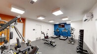 Located at 431 Church St in Norfolk, Virginia, this fitness center offers a well-organized, single-floor exercise space designed to support a well-rounded workout routine. The room is finished with a durable speckled rubber floor, providing both traction and impact absorption suited for high-activity use.

The space is divided into distinct functional zones. The cardio area features multiple treadmills, a stationary cycling bike, and an elliptical trainer, all positioned beneath bright recessed panel lighting that keeps the environment well-lit and energizing. A wall-mounted flat-screen television is positioned above a bold blue accent wall, adding an entertainment element to cardio sessions.

On the opposite side, a vibrant orange accent wall anchors the strength training zone, which includes a multi-function cable machine, an adjustable weight bench, and a full rack of dumbbells in graduated sizes. A large wall mirror expands the visual depth of this area and supports proper form during strength exercises. Climate control is provided by a wall-mounted mini-split unit, ensuring year-round comfort.

The overall design is clean and modern, with white walls and flush-mounted ceiling lights creating a bright, motivating atmosphere. The space is efficiently laid out to maximize utility while maintaining clear circulation paths between equipment zones.