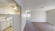 This single-floor residential unit offers a practical and well-organized layout designed for comfortable everyday living. The kitchen is outfitted with white cabinetry providing ample storage, light-toned quartz-style countertops, an electric coil range, and a refrigerator. Wood-look flooring grounds the kitchen area, lending a clean and modern feel. Adjacent to the kitchen, a hallway connects the main living spaces, with smooth neutral-painted walls and textured ceilings running throughout the unit. The living area is generously sized, featuring wall-to-wall carpeting in a warm taupe hue and a large vertical-blind window treatment that allows natural light to fill the room while offering privacy. A bedroom continues the carpeted flooring and includes a window with exterior views, providing additional natural light. The bathroom is efficiently designed with a combined tub and shower unit, a toilet, a vanity, and wood-look tile flooring that complements the rest of the unit's finishes. Clean white walls and simple fixtures give the bathroom a bright, functional character. Overall, the unit presents a cohesive, neutral palette with a straightforward floor plan that maximizes usable space across each room.