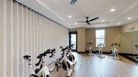 Core Fitness is a thoughtfully designed fitness center that offers a clean, modern environment for effective workouts. The space features hardwood-style flooring throughout, providing a durable and polished aesthetic that complements the neutral, warm-toned walls. A tray ceiling adds architectural character to the room, while recessed lighting and a sleek ceiling fan ensure the space remains well-lit and comfortably ventilated during exercise sessions. Large windows along the perimeter flood the interior with natural light and provide views of the surrounding area, creating an open and energizing atmosphere. The fitness floor is outfitted with a row of stationary cycling bikes, ideal for high-intensity cardio training, along with additional cardio and strength equipment positioned near the windows to take full advantage of the natural light. Vertical blinds along one wall offer flexible light control and privacy as needed. The layout is spacious and functional, allowing for free movement between equipment. The overall ambiance is clean, bright, and motivating — well-suited for both focused individual workouts and group cycling sessions.