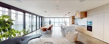 This 1,700-square-foot amenity floor is a sophisticated, high-rise retreat designed to deliver a premium resident experience. The space is unified by polished terrazzo flooring throughout, which reflects abundant natural light and lends a refined, contemporary aesthetic. A glass-lined corridor runs along the perimeter, offering unobstructed views of the outdoor pool deck, complete with lounge chairs positioned to take in sweeping panoramic vistas of the surrounding cityscape and horizon.

The central lounge area is anchored by an expansive sectional sofa, a wall-mounted flat-screen television, and a sleek linear fireplace set against a dramatic marble stone surround. Floor-to-ceiling windows frame striking high-rise city views on multiple sides, creating an immersive urban backdrop. A unique suspended wicker hanging chair and built-in shelving add character and visual interest to the lounge.

The full catering kitchen is appointed with light wood cabinetry, a large marble waterfall island with bar seating, a full marble backsplash, integrated appliances including double wall ovens, and a gas cooktop — designed to support events and social gatherings. Additional seating areas with designer chairs and indoor greenery provide intimate spaces throughout. Concrete accent walls and warm wood paneling complete the elevated, modern-minimalist design language of this exceptional amenity space.