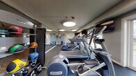 Located at 3316 Foxboro Dr in Woodridge, Illinois, this fitness center is a thoughtfully designed, single-floor facility offering a comprehensive workout environment. The main exercise area is outfitted with a row of Precor treadmills and elliptical trainers positioned along rubber flooring, providing a stable, impact-absorbing surface suited for high-intensity use. Large windows along one wall allow natural light to fill the space, offering views of the surrounding outdoor greenery. Wall-mounted flat-screen televisions are strategically placed throughout, keeping users engaged during cardio sessions. Full-length mirrors line one wall, enhancing the sense of openness and allowing users to monitor their form. A dedicated free weights section includes a dumbbell rack and weight bench, while a shelved storage unit holds medicine balls and additional accessories for functional training. A cable machine is also visible in the mirrored section, accommodating strength and resistance workouts. Adjacent to the main floor, a glass-enclosed studio area features hardwood-style flooring and colorful tile accents, suitable for stretching, yoga, or small group sessions. Flush circular ceiling lights provide consistent, even illumination throughout. The overall ambiance is clean, modern, and functional.